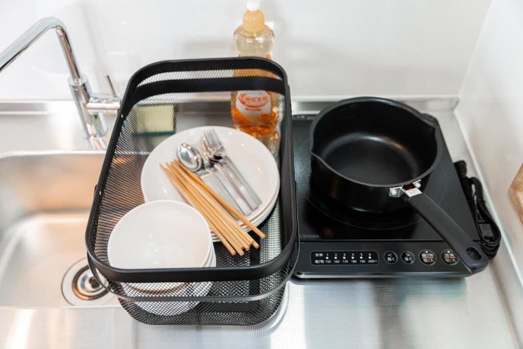 A kitchen counter with a portable induction cooktop, a black frying pan, a bottle of cooking oil, a metal strainer, chopsticks, and a plate with utensils.