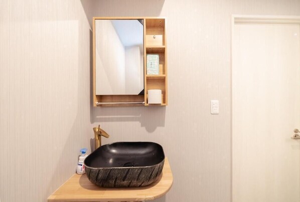 A bathroom with a black vessel sink, a wooden vanity, a mirror, and a small shelf with toiletries.