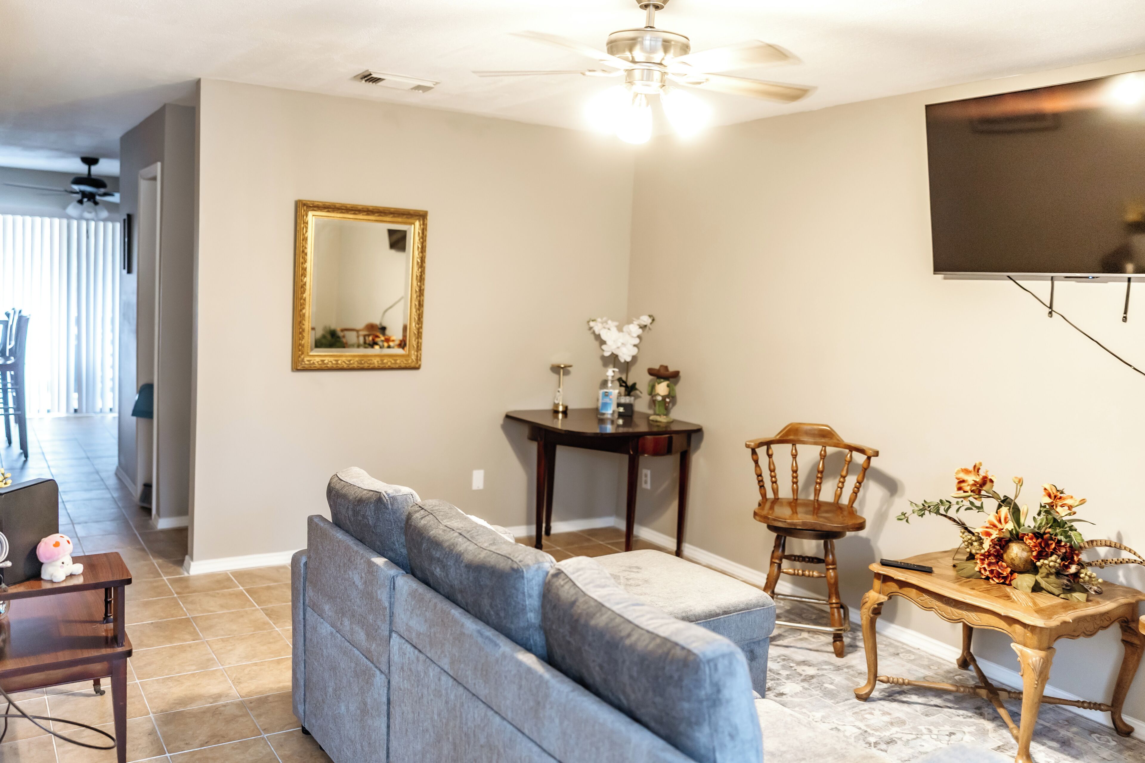 A living room with a sofa, a dining table with a vase of flowers, a wooden chair, a television mounted on the wall, and a ceiling fan.