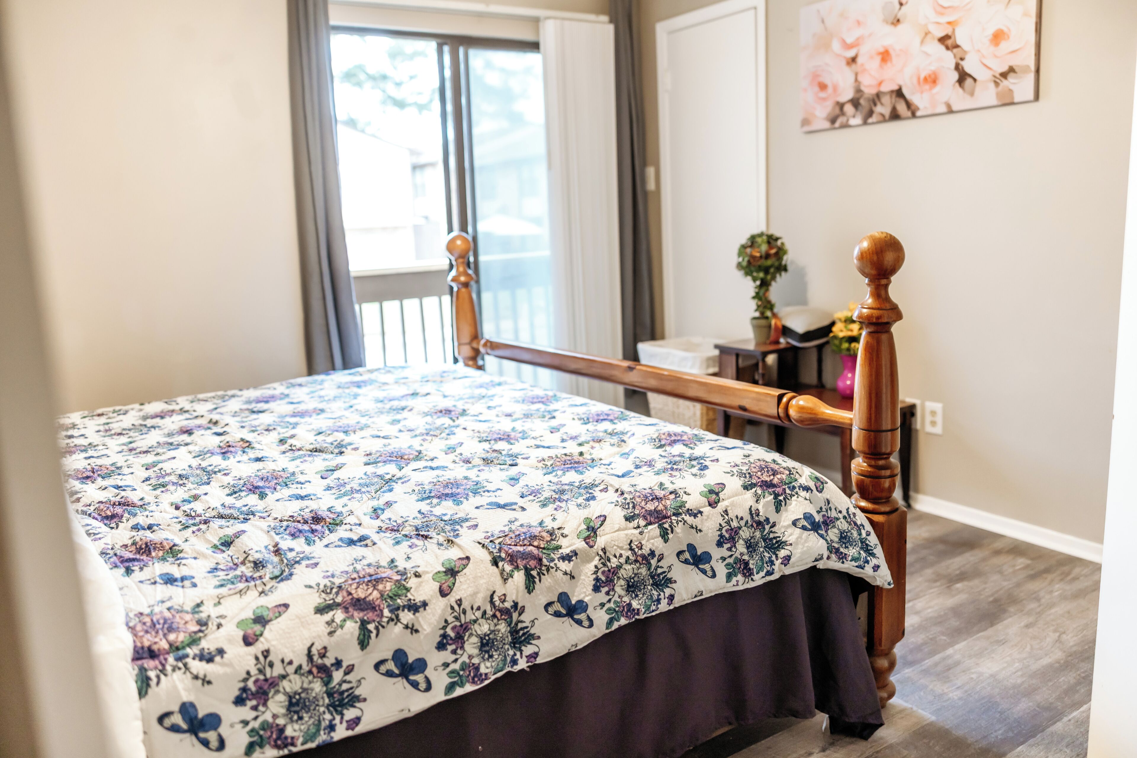 Une chambre à coucher avec un lit en bois, une literie à motifs floraux, une fenêtre avec des rideaux et un tableau accroché au mur.