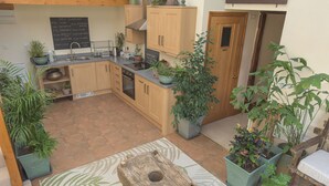 Une cuisine compacte avec des meubles en bois, un évier et un tableau noir affichant le programme quotidien. Plusieurs plantes en pot sont disposées dans l’espace.