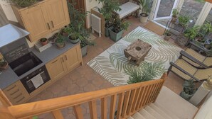 Une cuisine avec des armoires en bois, un four encastré et un plan de travail muni d’un distributeur de liquide vaisselle. On y trouve plusieurs plantes en pot sur le sol et un tabouret en bois au centre.