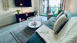 Uma sala de estar moderna com televisão de tela plana, uma mesa de centro de vidro, um sofá e vista para a cidade através de grandes janelas.