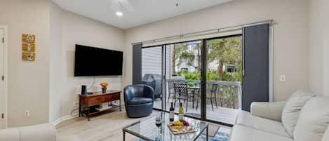 A modern living room with a flat-screen TV, leather sofa, glass coffee table, and a balcony with sliding glass doors.
