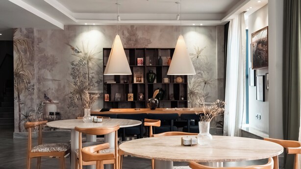 Uma área de refeições moderna com mesas e cadeiras redondas, iluminação pendente e uma parede decorativa com um mural floral.