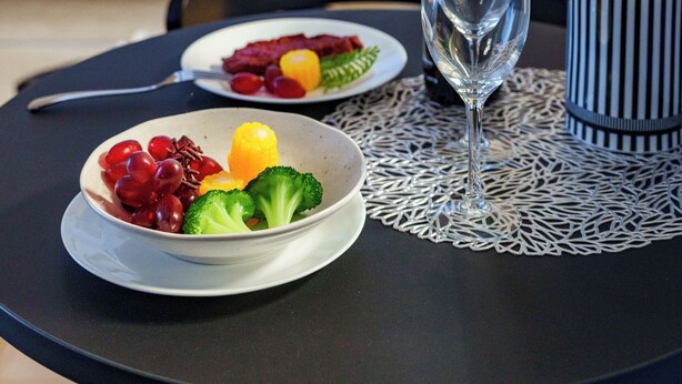 A dining table with a bottle of wine, a glass, and a bowl of fruit and vegetables. A bed with white bedding is visible in the background. - E-US NAEBO STATION 103 (Sapporo)