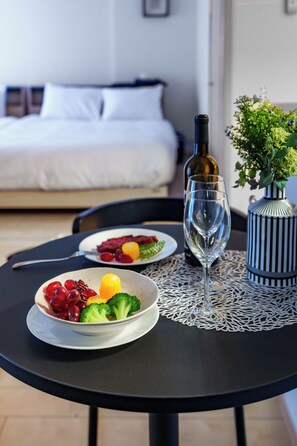 Een eettafel met een fles wijn, een glas en een schaal fruit en groenten. In de achtergrond is een bed met wit beddengoed te zien.