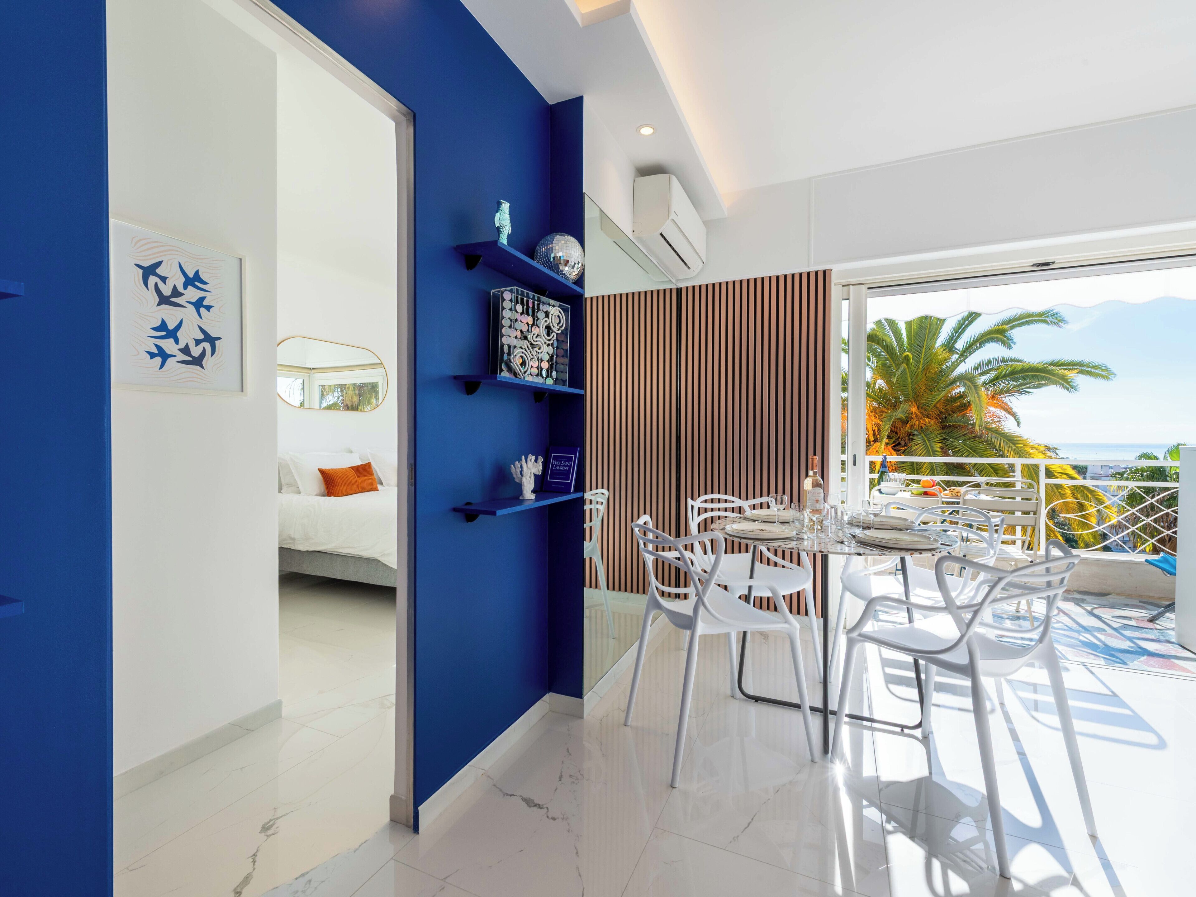 A modern living space with a blue accent wall, a dining area with white chairs, and a view of the ocean.