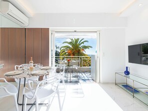 A modern dining area with a table set for four, white chairs, and a view of palm trees and the sea through an open door. - 2-Br Sea-View Apartment, Residence with Swimming Pool – Cannes (Cannes)