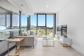 A modern living room with a sofa, coffee table, and TV, large windows with a city view, and a balcony. - City Heart 1-Bedroom Steps from Lake with Parking (Canberra)