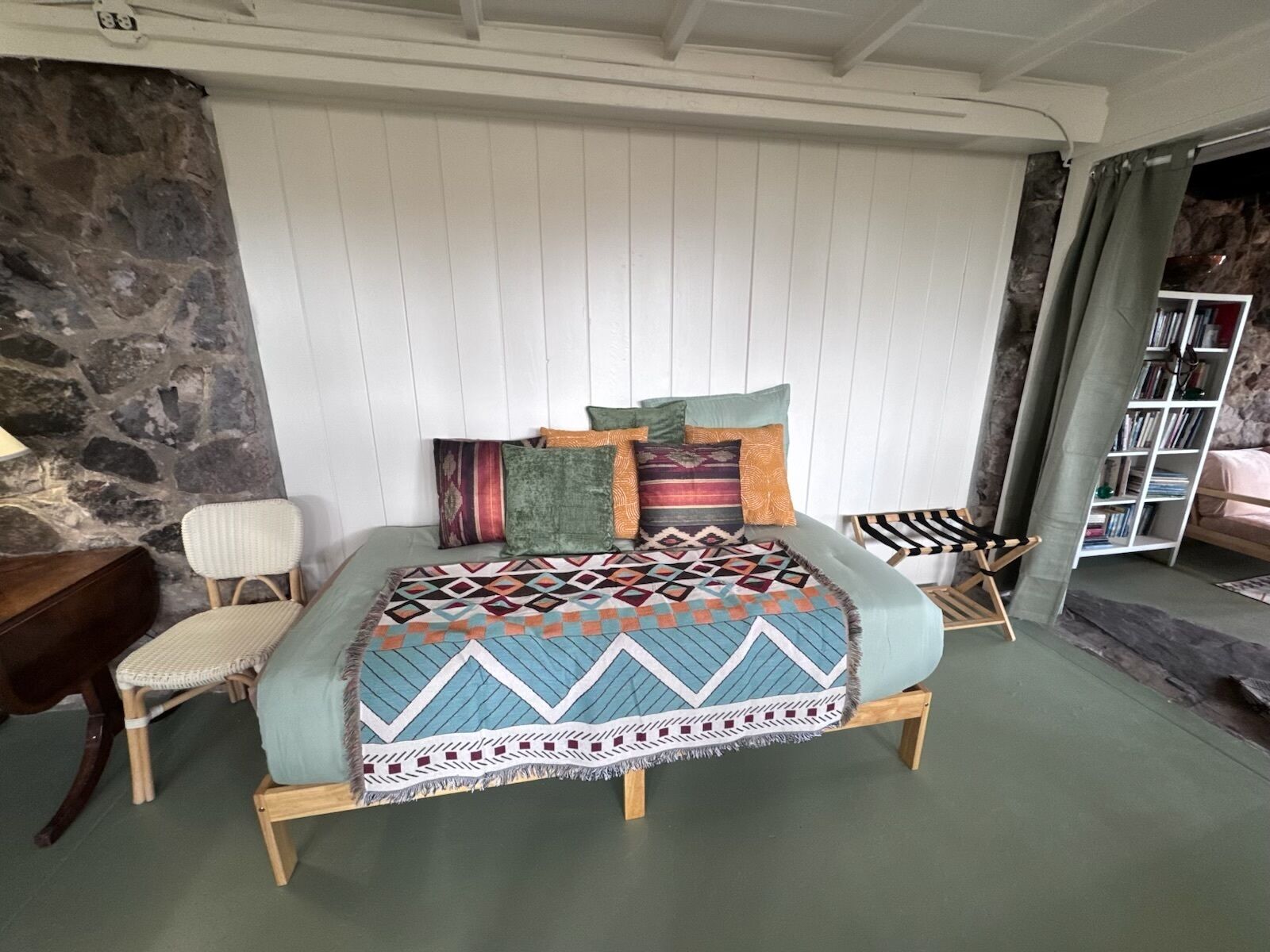 A bed with a patterned bedspread, a wooden headboard, and multiple pillows. There is a chair and a small table beside the bed.