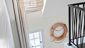 A room with a sloped ceiling, a hanging woven light fixture, a window with curtains, a wall-mounted circular object, and a staircase with black railings. - New Crow’s Nest Ocean View l Downtown l Queen Suite (Newburyport)