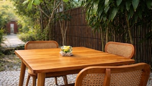 A wooden table with a glass bowl containing yellow flowers, surrounded by wicker chairs, set on a gravel path with a bamboo fence in the background. - Dapur Alit Villas (Kasihan)