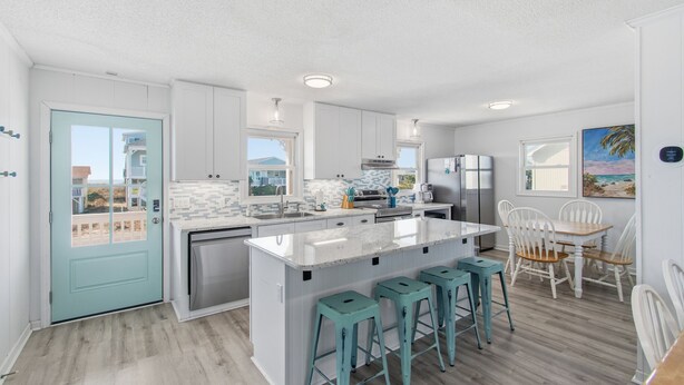 A modern kitchen with white cabinets, a central island with bar stools, and a dining area with white chairs. - "Once in a Blue Moon - 796 OBW" 4 Bedroom, Second Row (Supply)