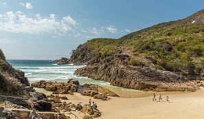 Ein felsiger Strand mit brechenden Wellen, umgeben von Hügeln und spärlicher Vegetation.