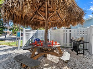 A wooden picnic table with a thatched umbrella, a grill, and picnic supplies. - Holiday House | Steps to Siesta Beach, Pet Friendly, Walk to Village (Siesta Key)