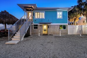 A two-story house with a staircase leading to the entrance, a thatched roof structure to the left, and a white fence surrounding the property. - Holiday House | Steps to Siesta Beach, Pet Friendly, Walk to Village (Siesta Key)