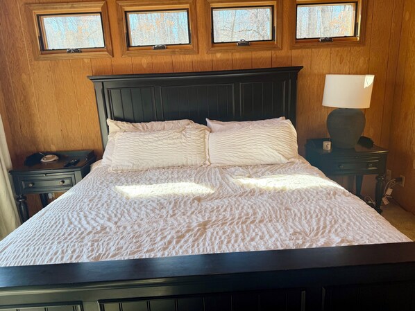 A neatly made bed with a dark headboard, a bedside table with a lamp, and three windows with a view of bare trees. - Woodland Retreat with a Swim Spa - Innsbrook Resort (Innsbrook)