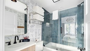 Ein modernes Badezimmer mit einem weißen Waschbecken, einem schwarzen Wasserhahn und einem Spiegel darüber. Eine Duschkabine aus Glas mit einem weißen Handtuch an der Tür.