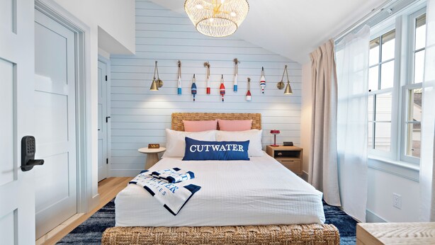 Ein Schlafzimmer mit Bett, Nachttisch, Wandlampe, Fenster mit Vorhängen sowie einer Wand mit maritimen Dekorationen.