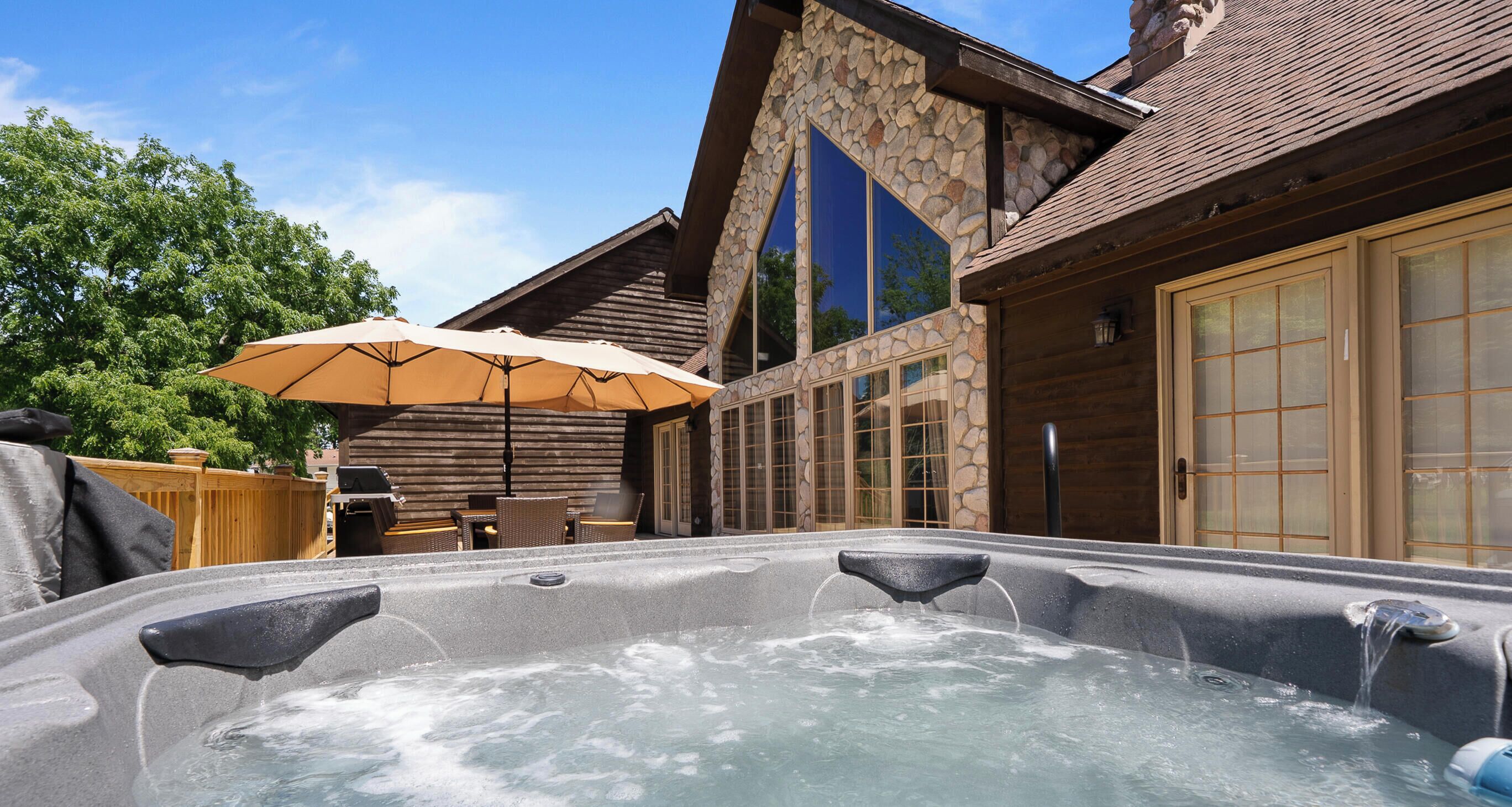A hot tub with a stone wall and wooden deck, a covered seating area, and a large window.
