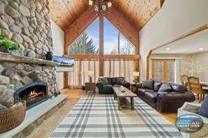 Ein gemütliches Wohnzimmer mit Kamin aus Naturstein, Ledersofas und einem großen Fenster mit Blick auf Bäume.