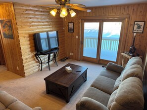 Interior de una cabaña de troncos con televisor, sofá y vista a un paisaje nevado a través de la puerta corrediza de vidrio.