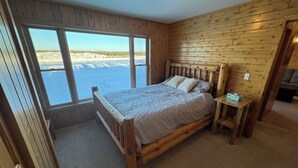 Ett sovrum i en stuga med träpanel, ett stort fönster, en säng, ett nattduksbord och utsikt över ett snötäckt landskap.