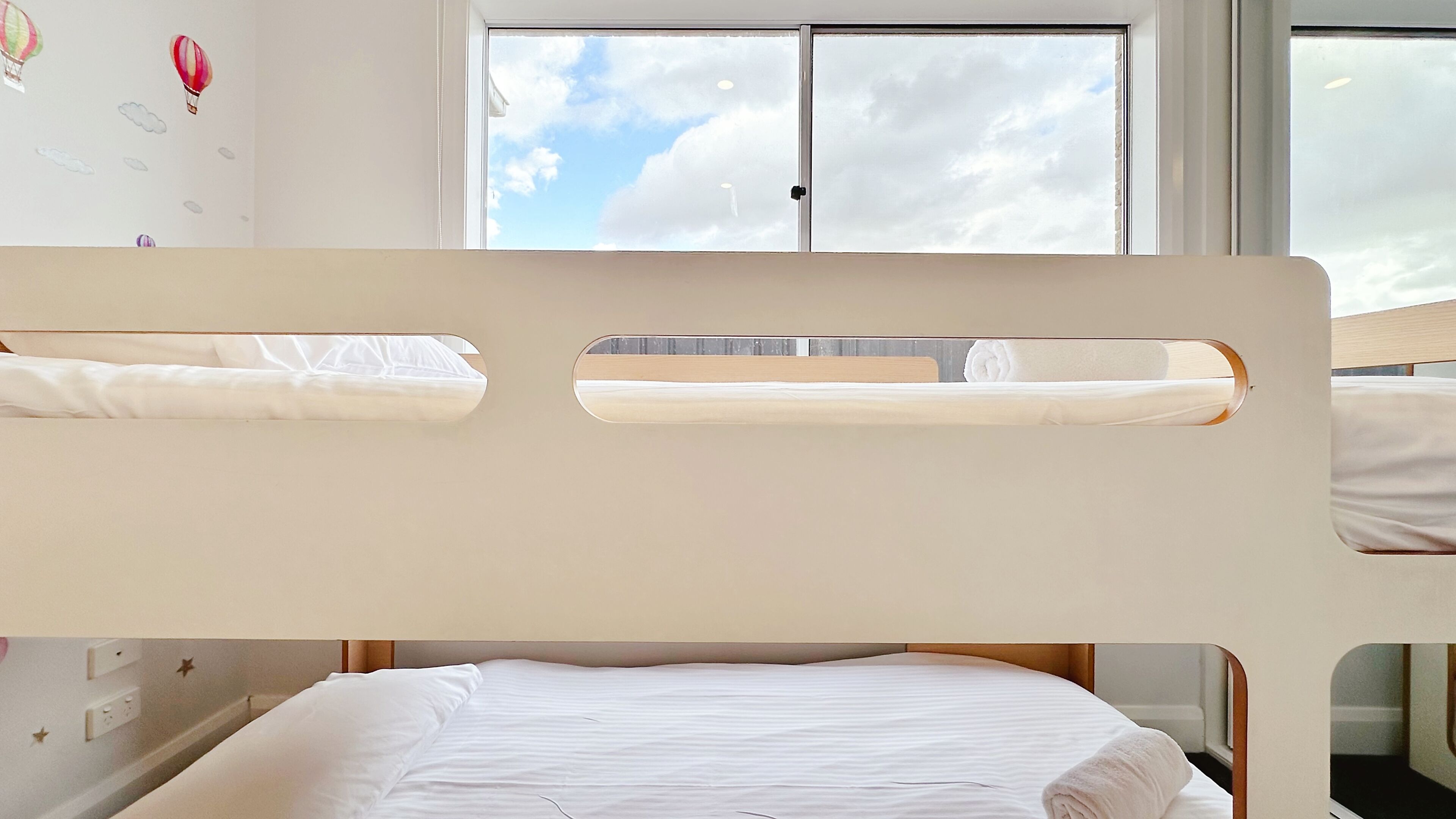 Un lit superposé avec une fenêtre, un lit recouvert d’une couverture blanche enroulée, et un mur décoré avec un motif de nuage et de montgolfière.