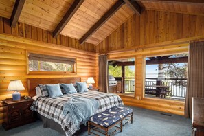 A log cabin bedroom with a bed, two bedside lamps, a bench, a view of the outdoors, and a grill outside.