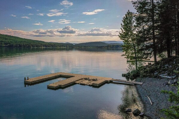 A calm lake with a wooden dock extending into the water, surrounded by trees and a clear sky.
