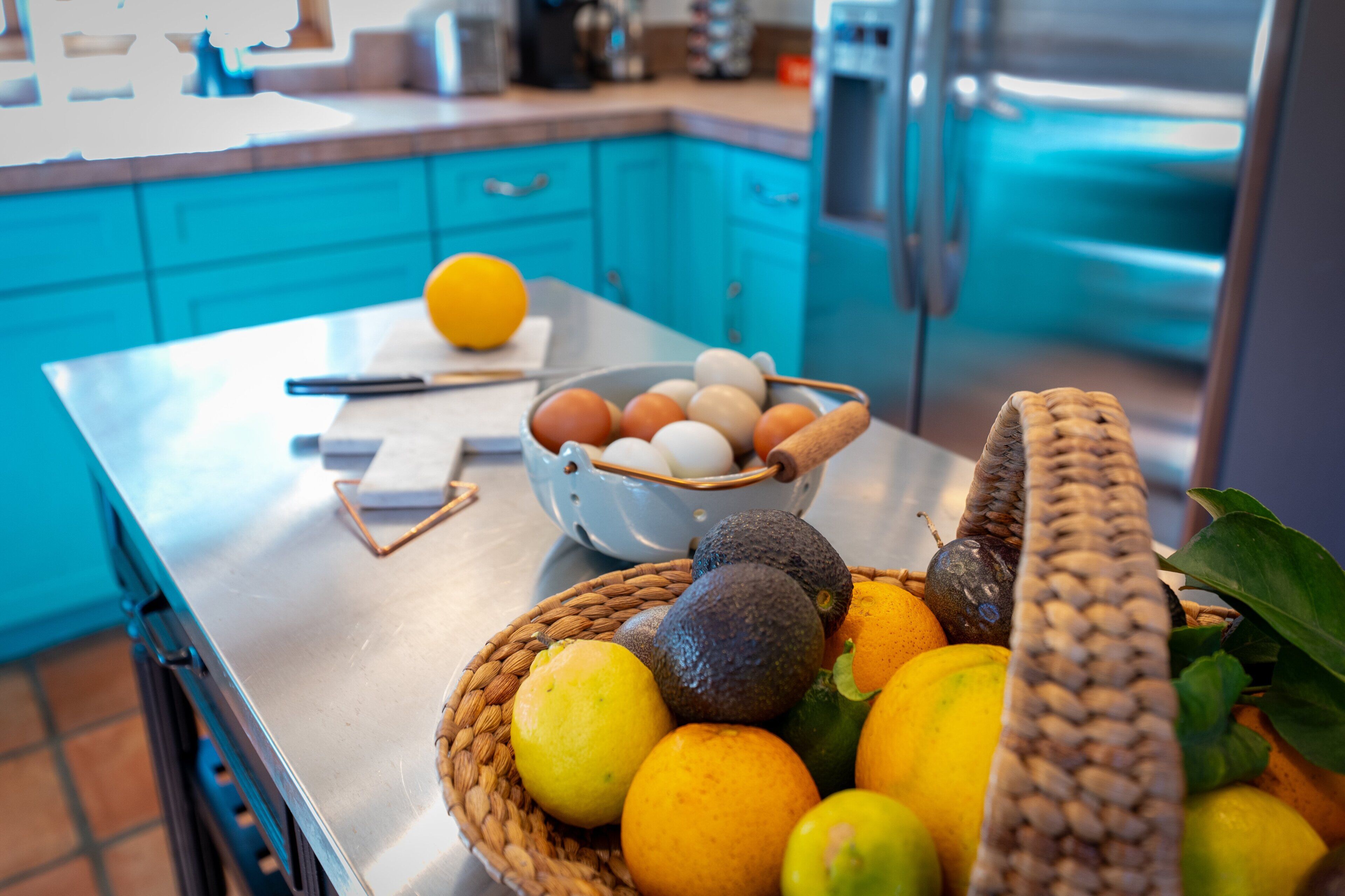 Uma bancada de cozinha com uma fruteira contendo laranjas, limões e abacates, e uma tigela de ovos.