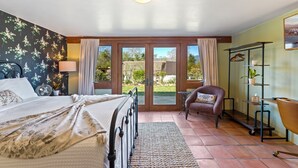 Ein Schlafzimmer mit Bett, Stuhl, Schreibtisch, einem Regal mit Pflanzen und Blick durch eine Glastür.