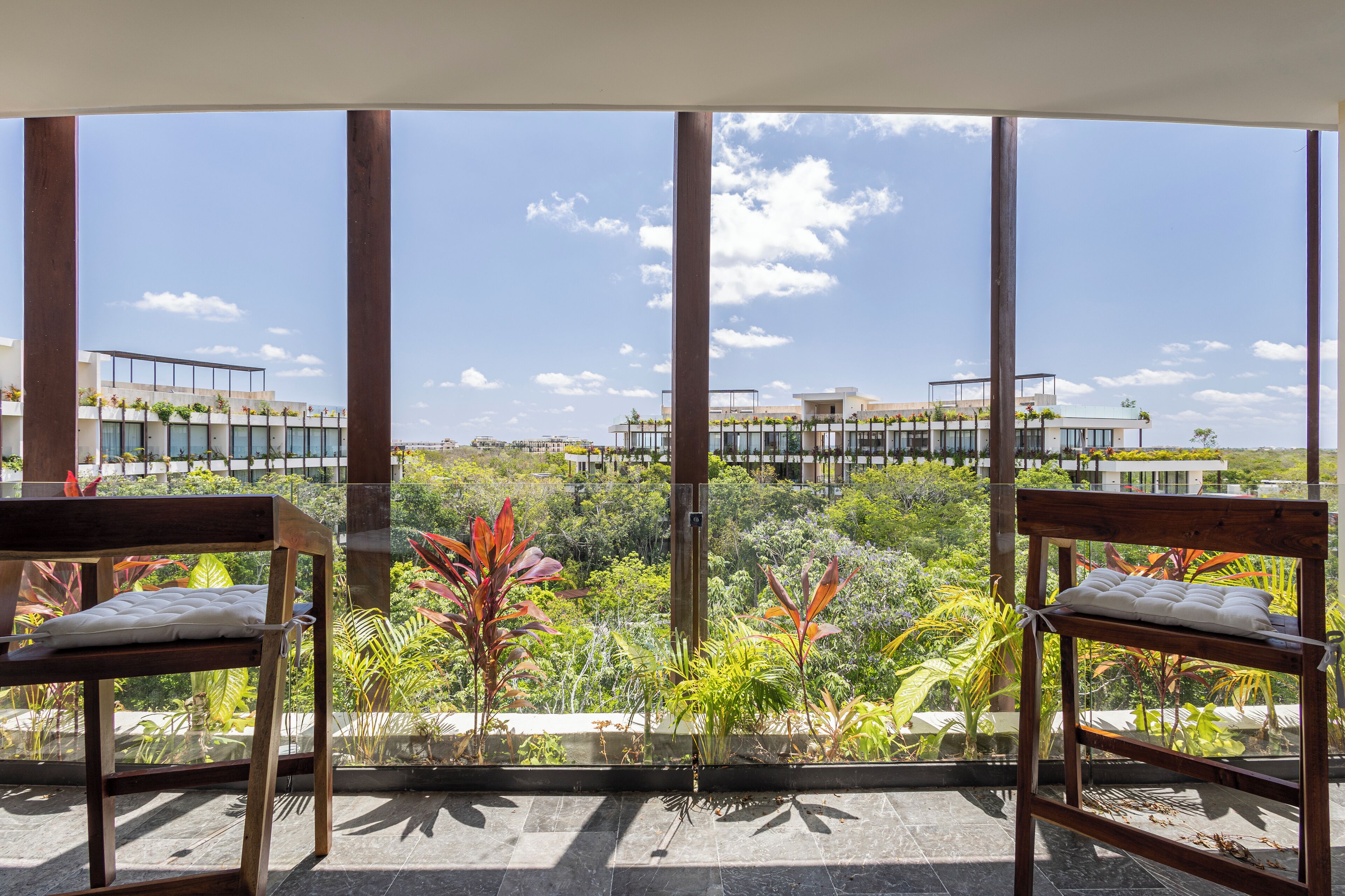 Ein Balkon mit zwei Holzstühlen, einer Glastrennwand und Blick auf ein modernes Gebäude sowie üppiges Grün.