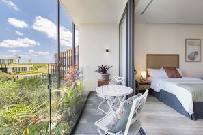A modern hotel room with a balcony, a bed, a bedside table with a lamp, and a view of greenery and a building outside. - Luxury Jungle Penthouse. Private Rooftop, Pool & High-Speed WiFi (Tulum)