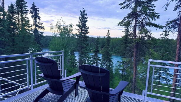 Two chairs on a balcony overlooking a forested area with a river. - Kenai Riverfront RV Camping & World Class Fishing. (Soldotna)