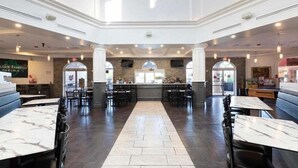 En rummelig restaurant med borde og stole med marmorplader, et centralt barområde og et højt loft med naturligt lys.