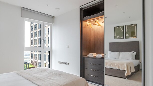A modern bedroom with a large bed, a walk-in closet, and a view of a building through a window. - Blueground | Whitechapel, fitness center, nr Tower of London (London)