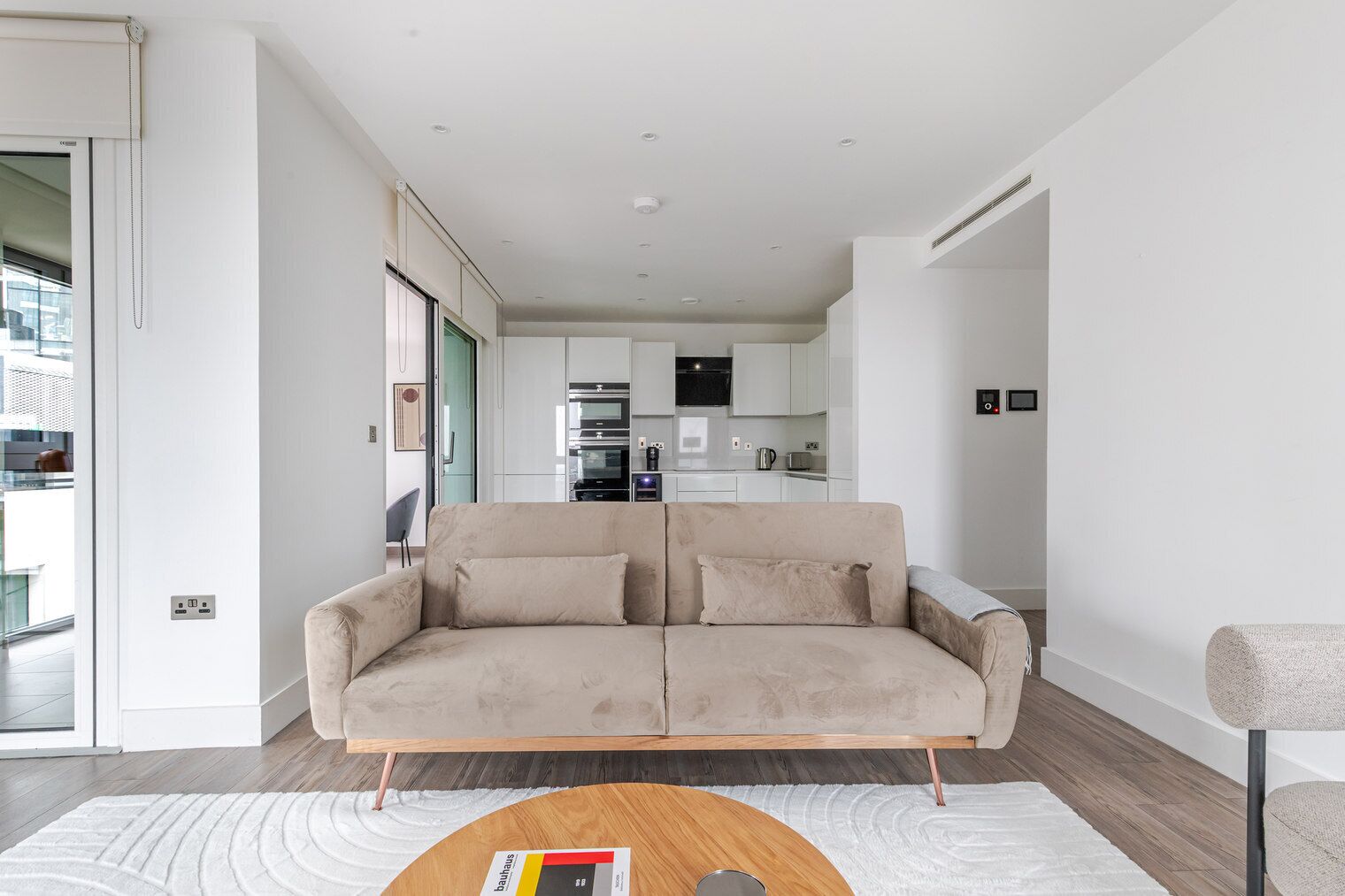 A modern living room with a beige sofa, a wooden coffee table, and a view of a kitchen and balcony.