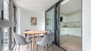 A modern dining area with a round table and four chairs, a kitchen visible through a glass door, and a city view through large windows. - Blueground | Whitechapel, fitness center, nr Tower of London (London)