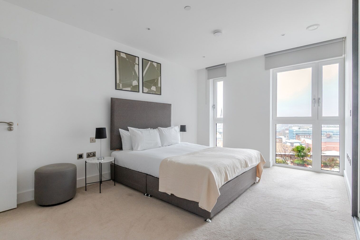 A modern bedroom with a large bed, a small side table, a lamp, and a view of the city through large windows.