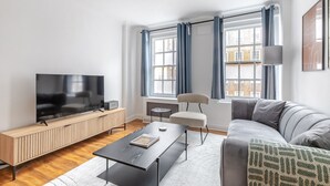 Ein modernes Wohnzimmer mit einem Flachbildfernseher auf einem Holz-TV-Möbel, einem grauen Sofa, einem kleinen runden Tisch und einem weißen Teppich.