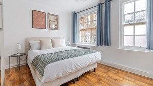 Ein Schlafzimmer mit Bett, Nachttisch, Fenster mit Vorhängen und Bildern an der Wand.