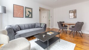 Ein modernes Wohnzimmer mit grauem Sofa, einem Couchtisch und einem Essbereich mit Stühlen und Tisch. An den Wänden befinden sich abstrakte Gemälde und eine Deckenleuchte.