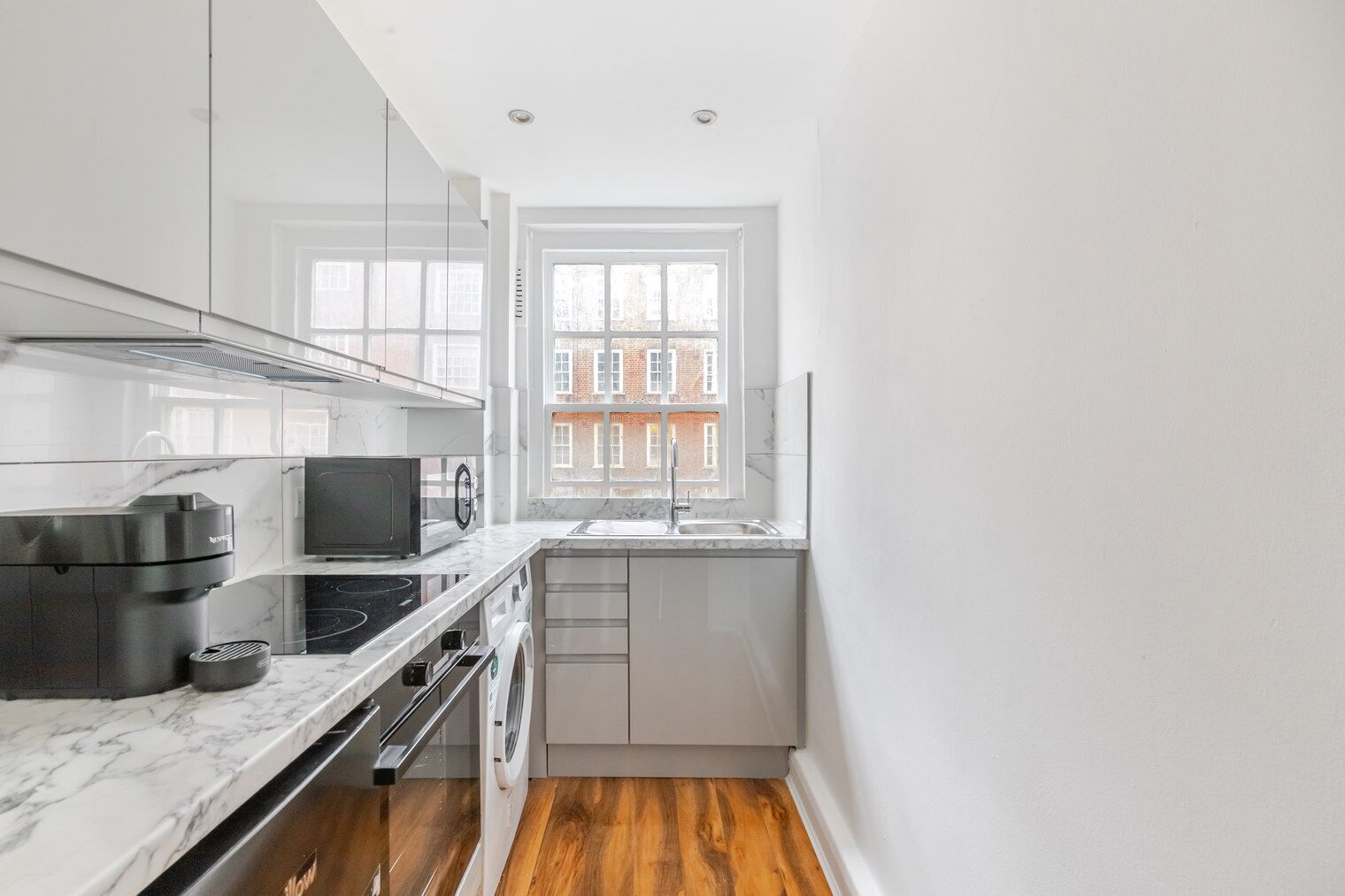 A compact kitchen with a built-in oven, microwave, and dishwasher, a window with a view of buildings, and a wooden floor.
