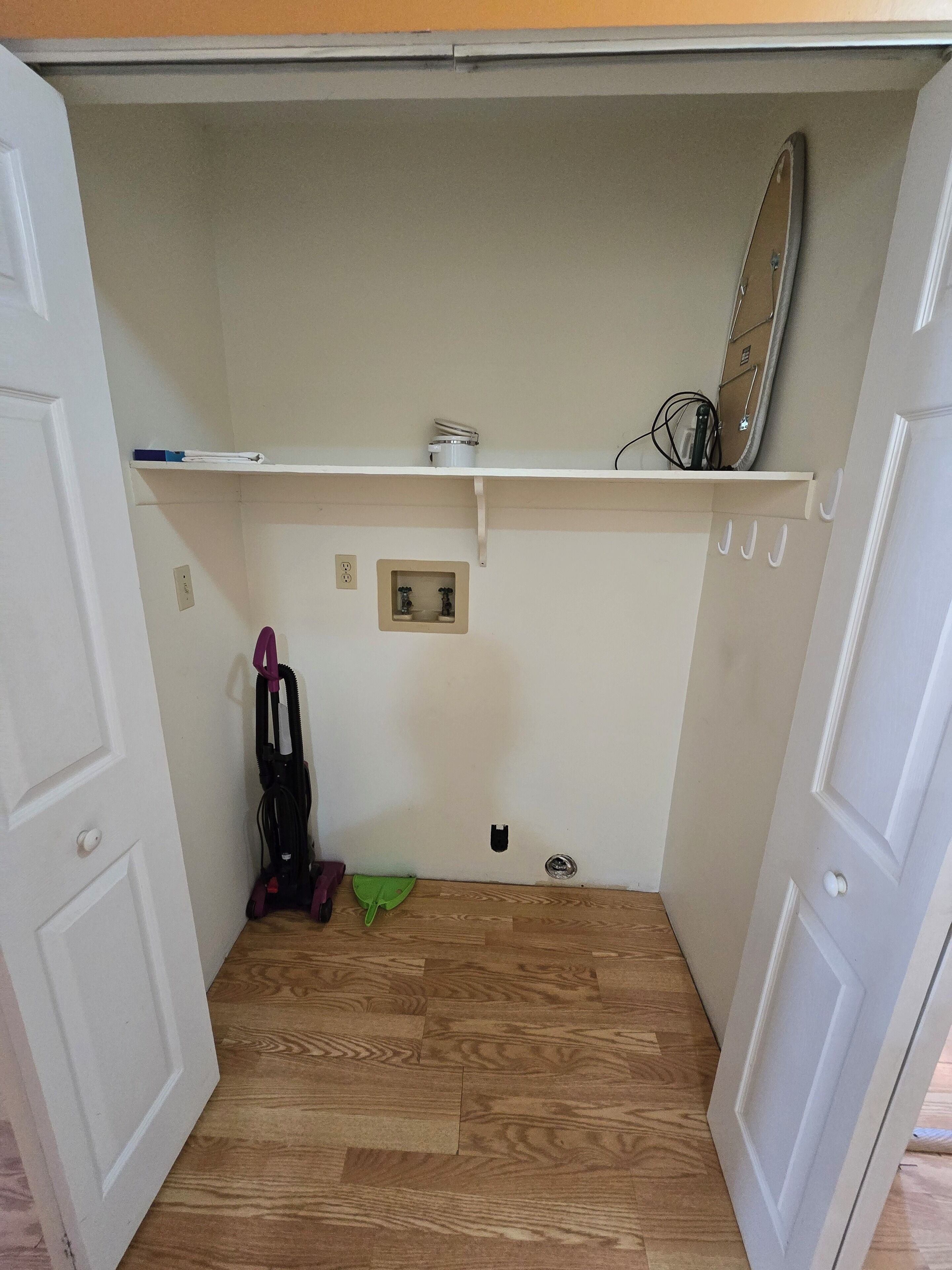 A compact room with a wooden floor, a white door, a small shelf with a mirror, and a vacuum cleaner.