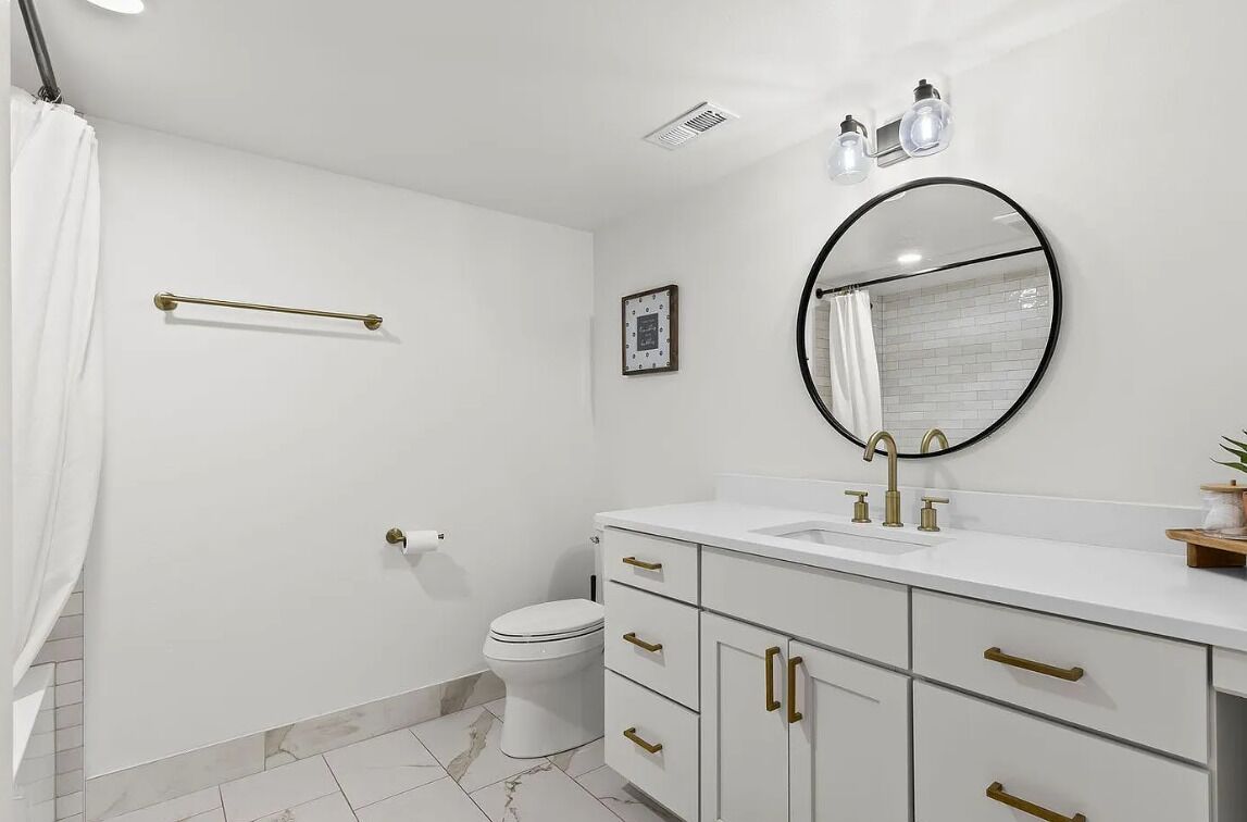 Een moderne badkamer met een wit wastafelmeubel, gouden handgrepen, een ronde spiegel en een toilet.