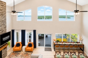 Un salon spacieux doté d’une cheminée, de deux fauteuils orange, d’un coin repas avec une table dressée pour six personnes, et de grandes fenêtres offrant une vue sur le lac.
