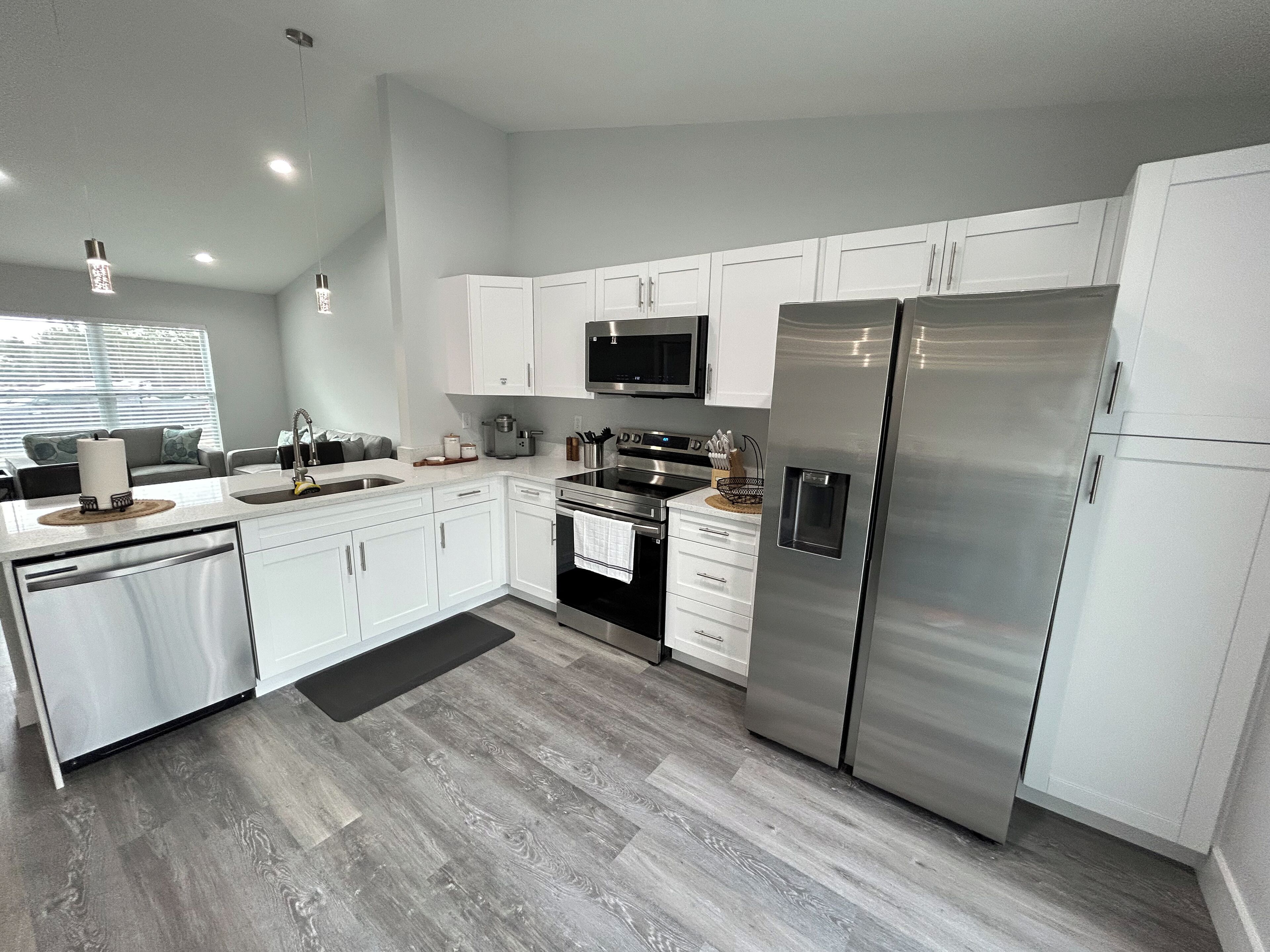 A modern kitchen with white cabinets, stainless steel appliances, and a grey wooden floor.