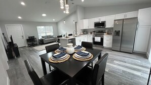A modern kitchen with stainless steel appliances, a dining area with a dark table and chairs, and a living area with a sofa and a TV. - 5.0 Rated 3BR Retreat • Gated Patio • Near WEC (OCALA)